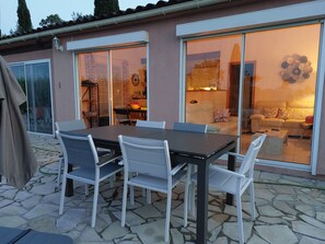 Une terrasse extérieure avec une table et des chaises, un espace salon couvert et une vue sur l’intérieur grâce à de grandes portes vitrées.