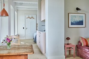 Un salon chaleureux avec une table en bois, un canapé rose et une petite table d’appoint. Une porte donne sur un coin cuisine équipé d’un meuble blanc et d’un luminaire suspendu.