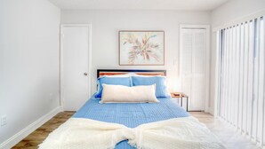 A bedroom with a bed, a nightstand, a white door, a window with blinds, and a wall-mounted painting.