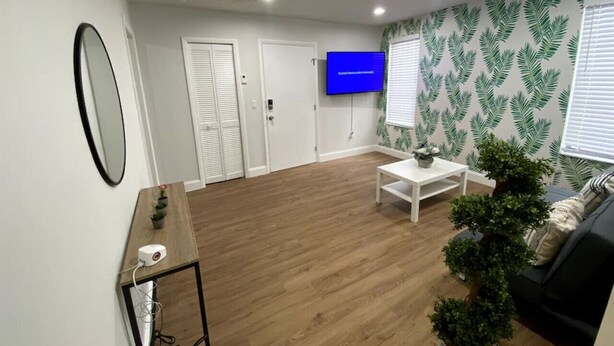 A modern living room with a wooden floor, a white sofa, a coffee table, a TV mounted on the wall, and a patterned wallpaper. - Renovated 2/1 apt w/ parking near Brickell Center (Miami)