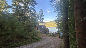 Een rustieke hut met een grindpad ernaartoe, omringd door dicht bos en een meer op de achtergrond.