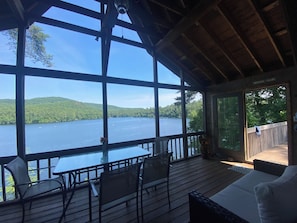 A covered deck with a table and chairs, overlooking a lake and forested hills.
