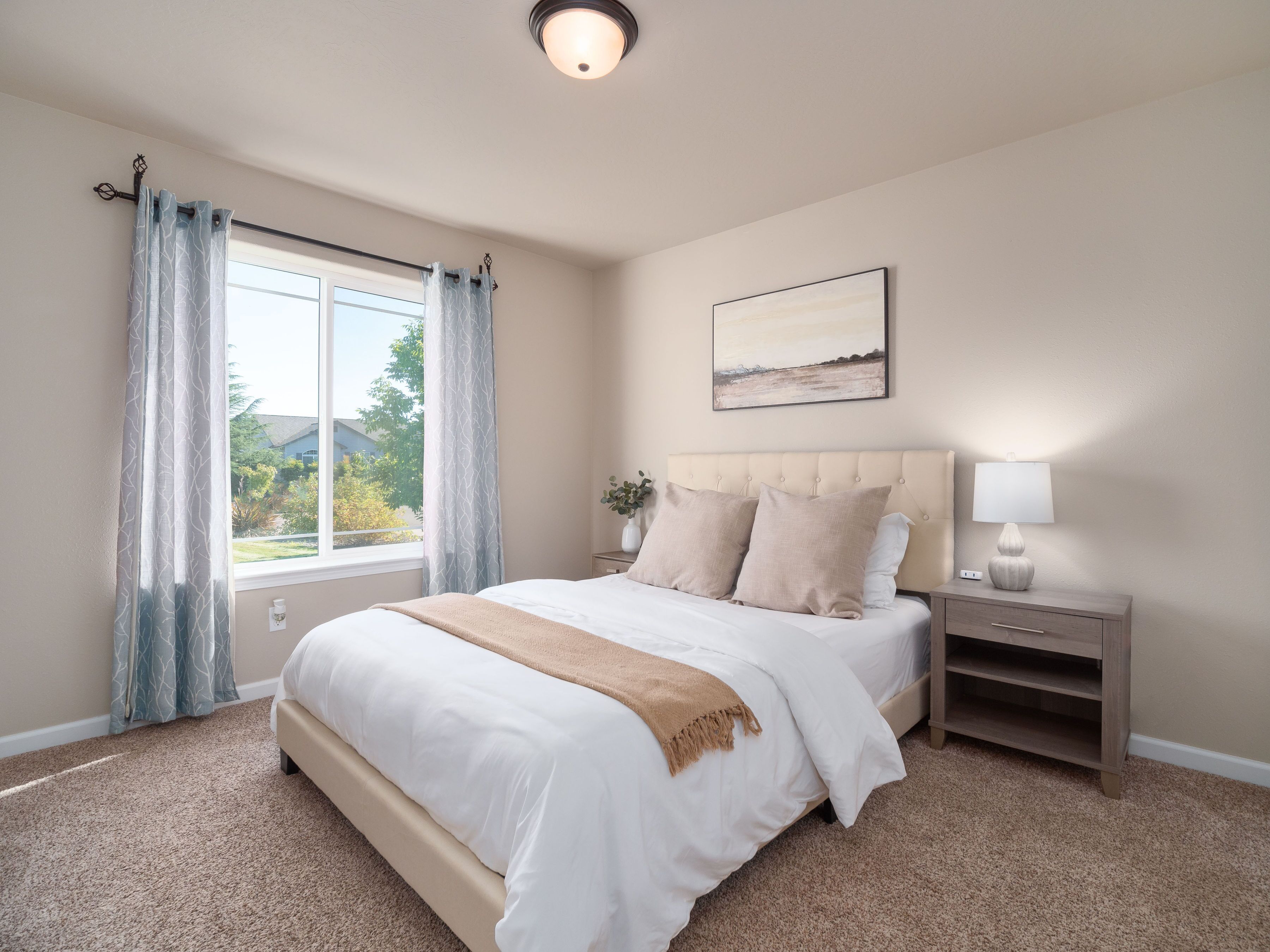 A bedroom with a large bed, a nightstand, a lamp, a window with curtains, and a painting on the wall.