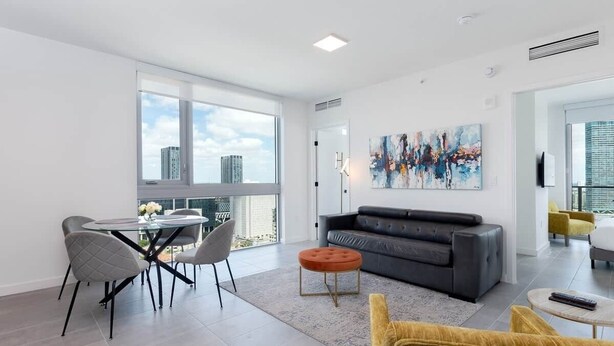 Un salon moderne avec un canapé, une table ronde et des chaises, une grande fenêtre donnant sur la ville et un tableau accroché au mur.