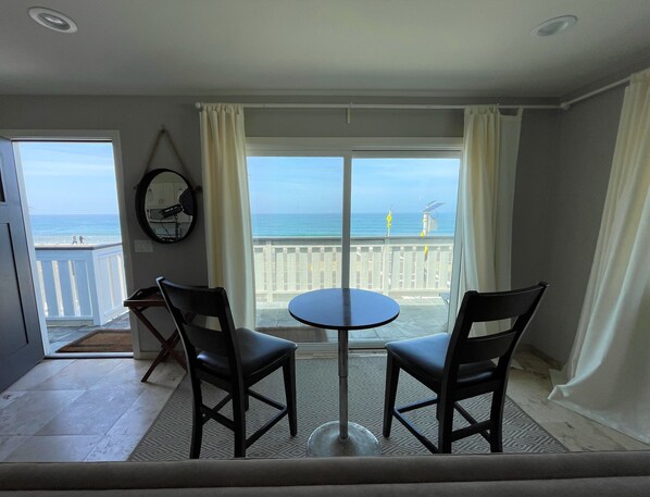 A room with a balcony overlooking the ocean, featuring a round table and two chairs, a ceiling fan, and a view of the sea.
