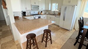 A modern kitchen with white cabinets, a granite countertop, a built-in oven, a microwave, a refrigerator, and two bar stools. - Beachside condo #B with 2bd /2 bath (Carlsbad)