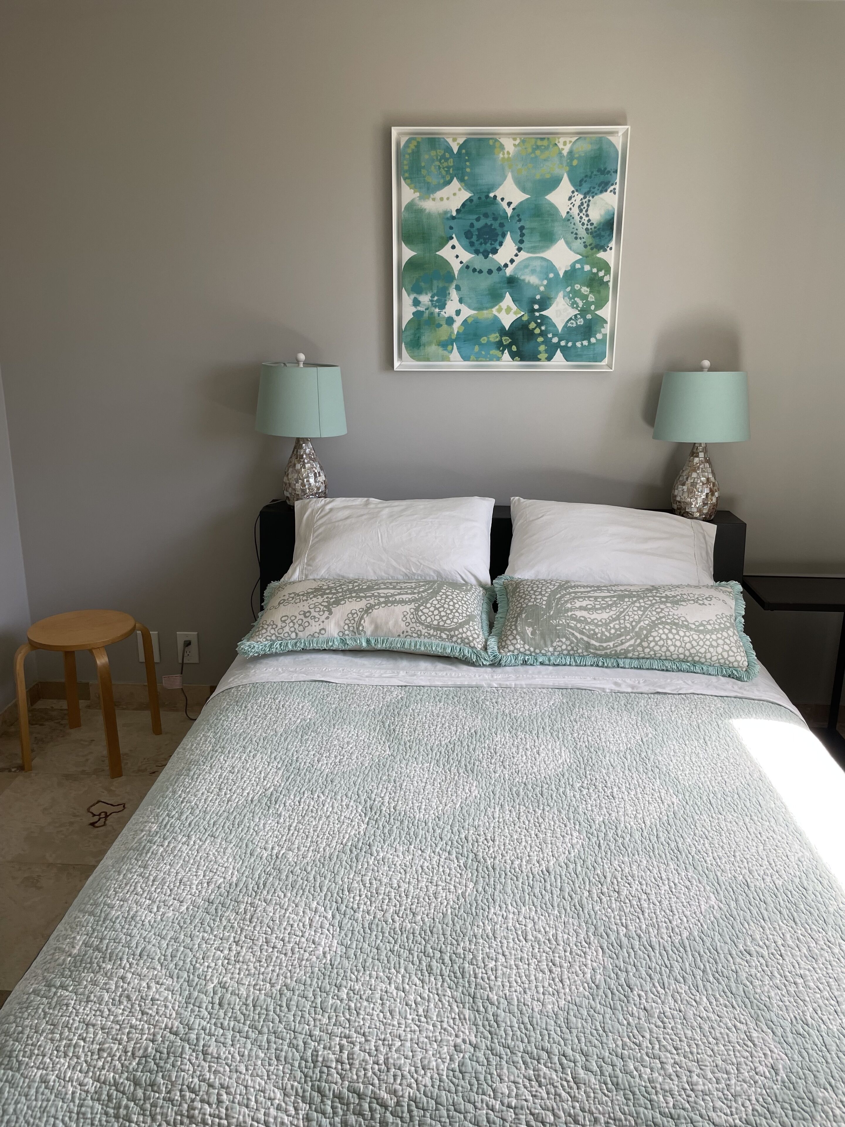A bedroom with a bed, two bedside lamps, a small stool, and a wall-mounted painting.