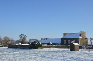 A snow-covered building with a brick exterior and a large chimney, surrounded by a fenced area and trees.