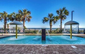 Piscina con fuentes de agua, rodeada de palmeras y un cielo despejado.
