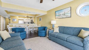 A living room with a blue sofa set, a dining area with white furniture, and a kitchen in the background. - Oceanfront Luxury 4BR Condo w/ Large Balcony & Pool (Myrtle Beach)