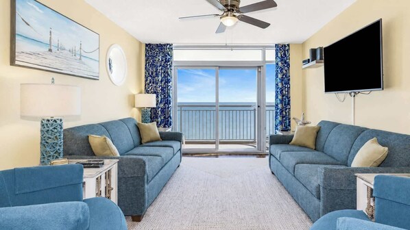 A room with two sofas facing a balcony, a ceiling fan, a flat-screen TV, and a painting on the wall.