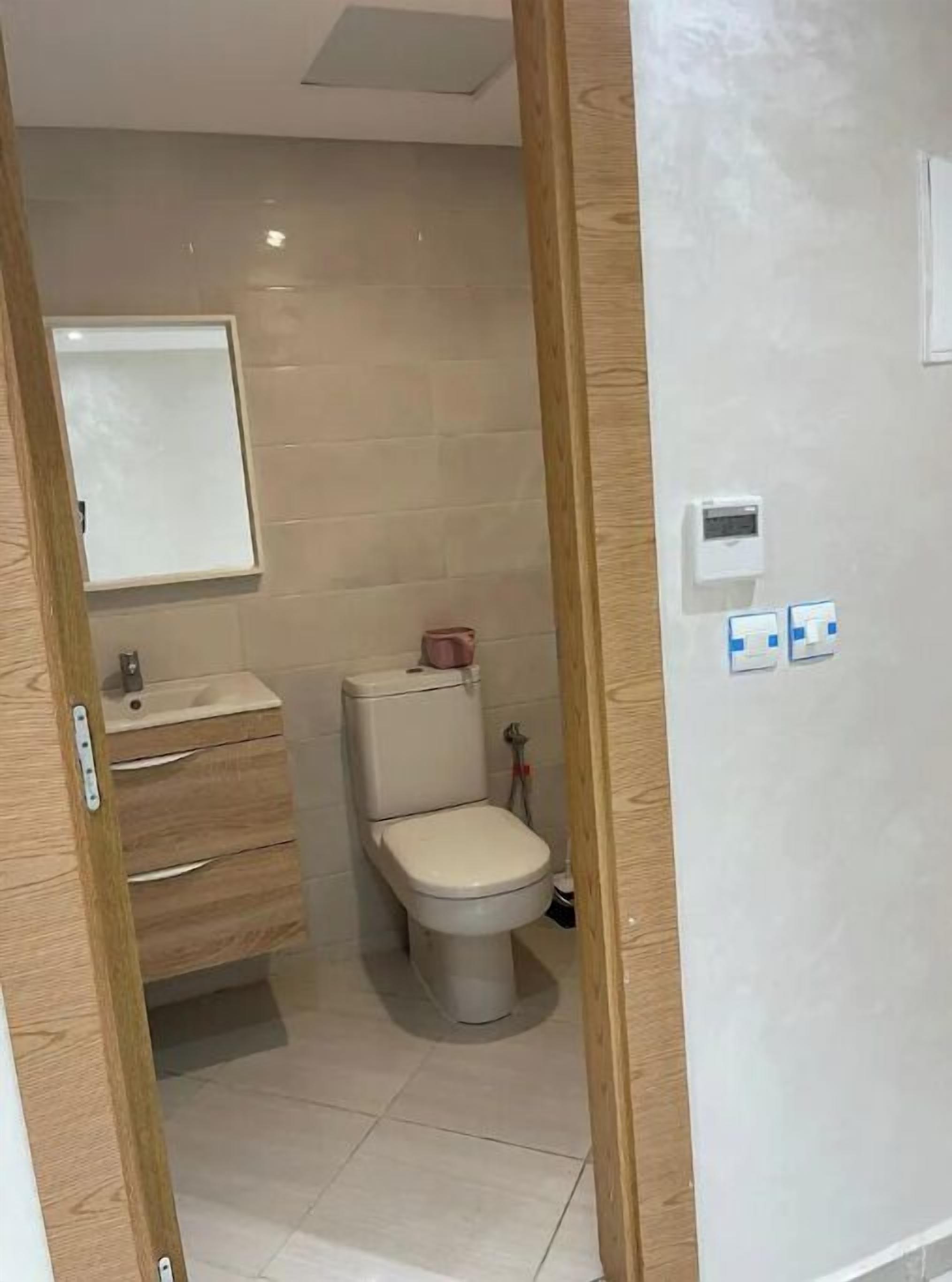 A small bathroom with a toilet, sink, and mirror. There is a wooden door frame and a wall-mounted electrical outlet.