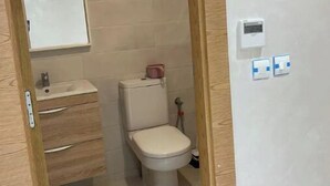 Baño pequeño con inodoro, lavamanos y espejo. Se observa un marco de puerta de madera y un enchufe eléctrico en la pared.