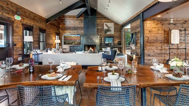 A spacious dining area with a long wooden table set for a meal, a fireplace, and a modern kitchen in the background.