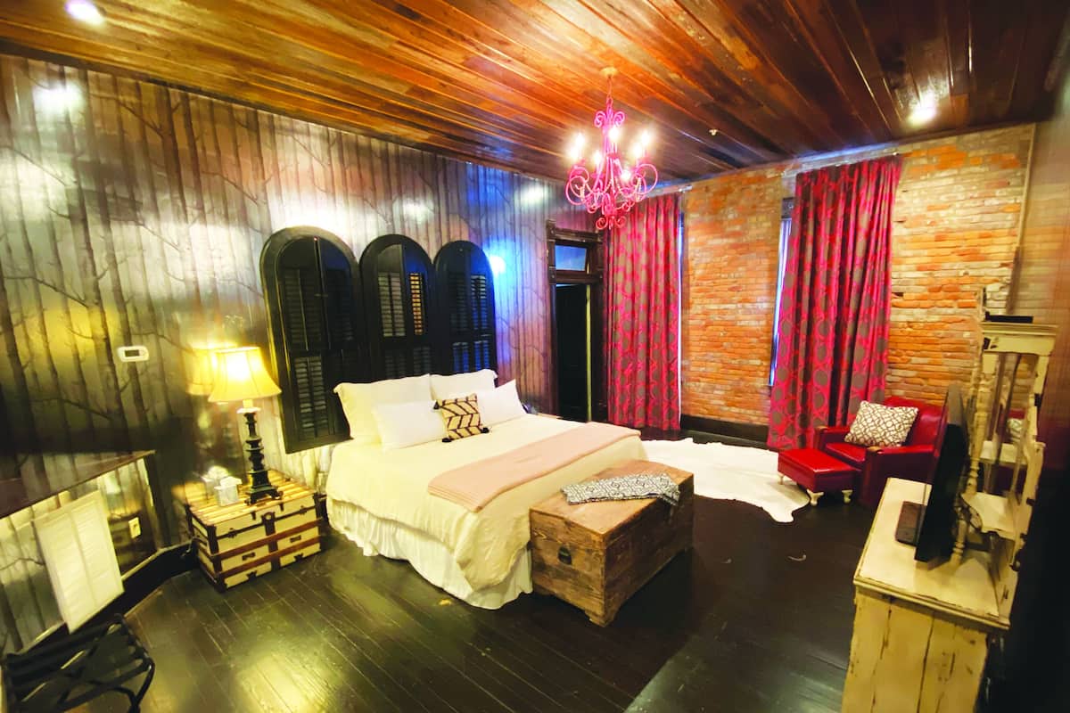 A bedroom with a large bed, wooden headboard, a chandelier, a red chair, and a wooden chest.