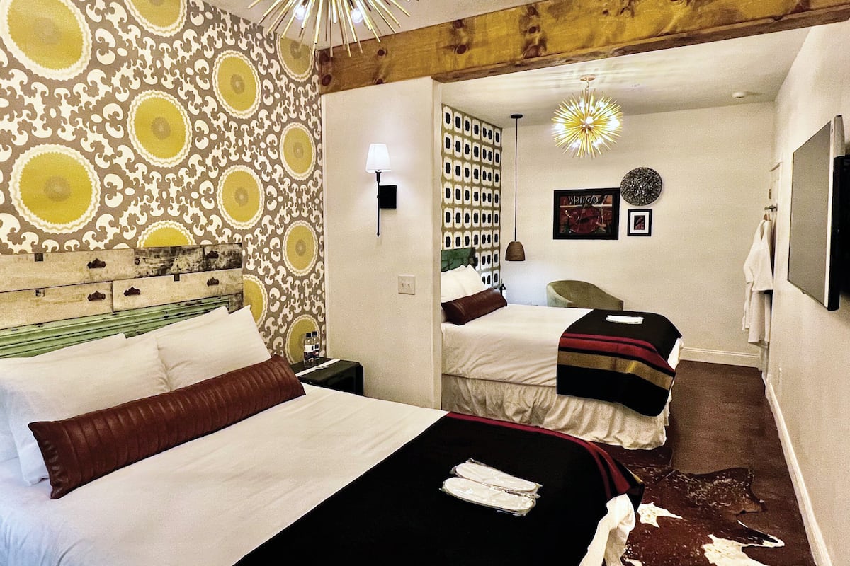 A hotel room with a patterned wallpaper, a bed with white linens, a headboard with a rustic design, a bedside table, a nightstand, a mirror, and a wall-mounted light fixture.