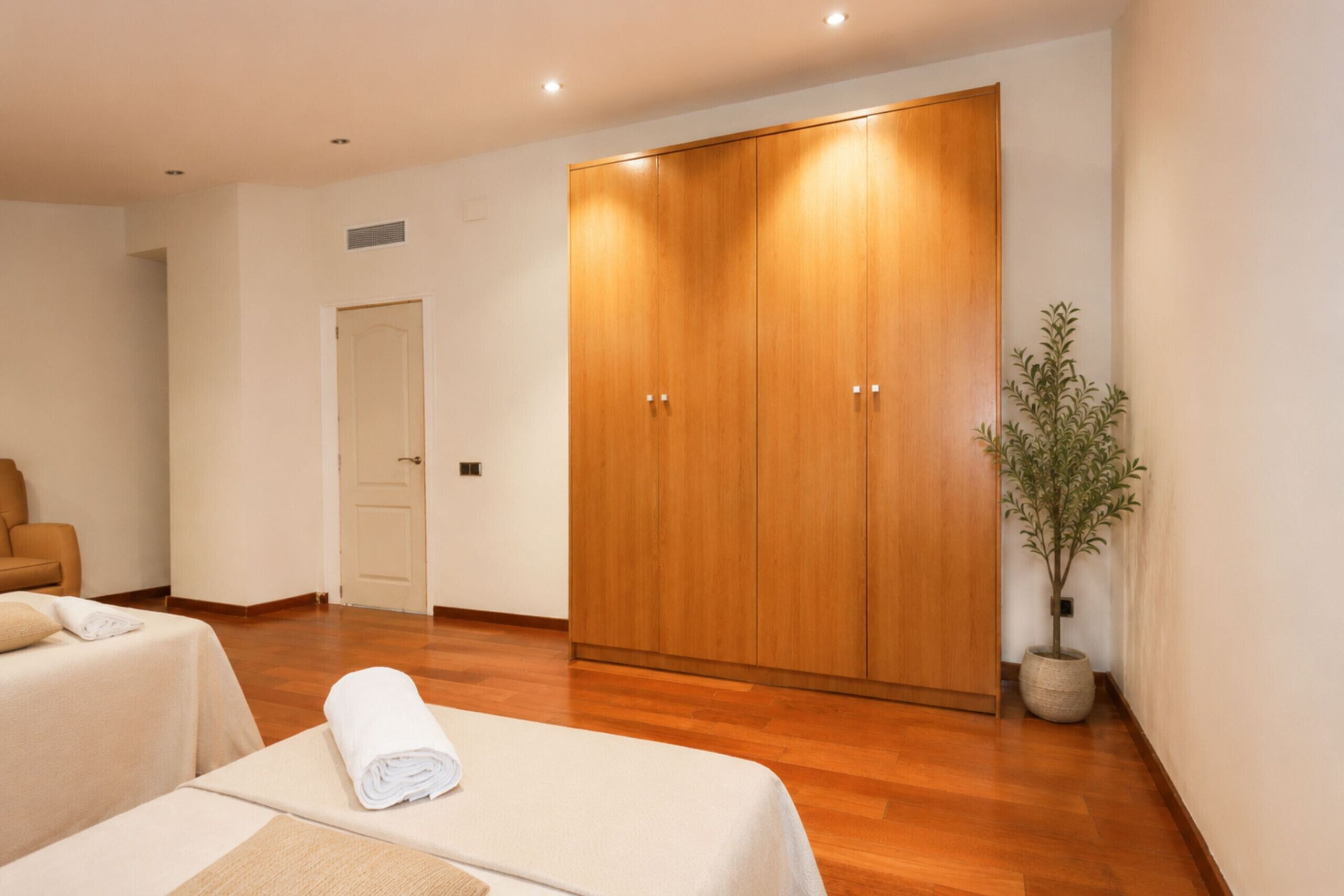 Hotelkamer met een houten garderobe, een bed met een opgerolde witte handdoek, een potplant en een houten vloer.