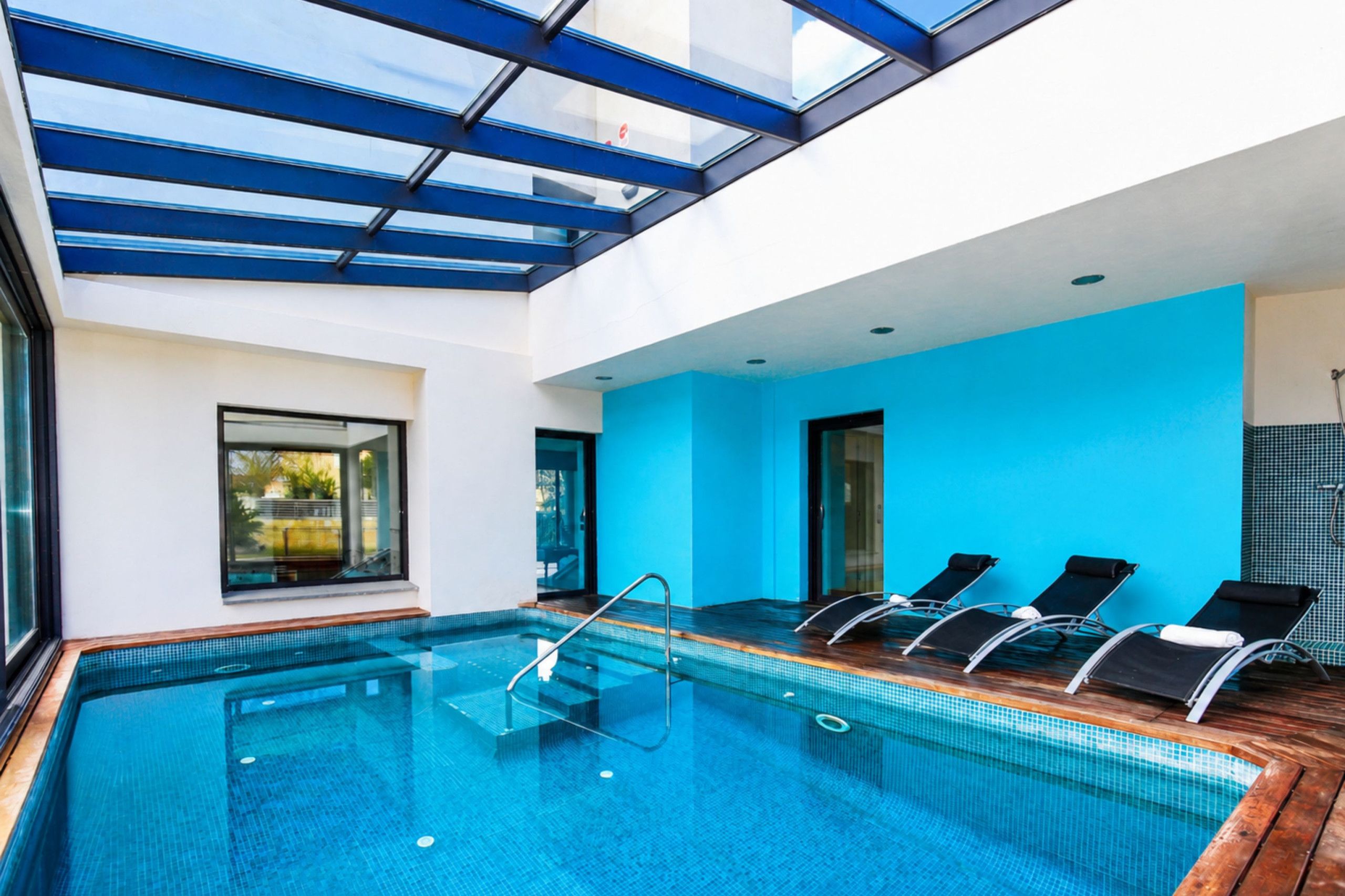 A modern indoor swimming pool with clear glass ceilings, blue walls, and lounge chairs.