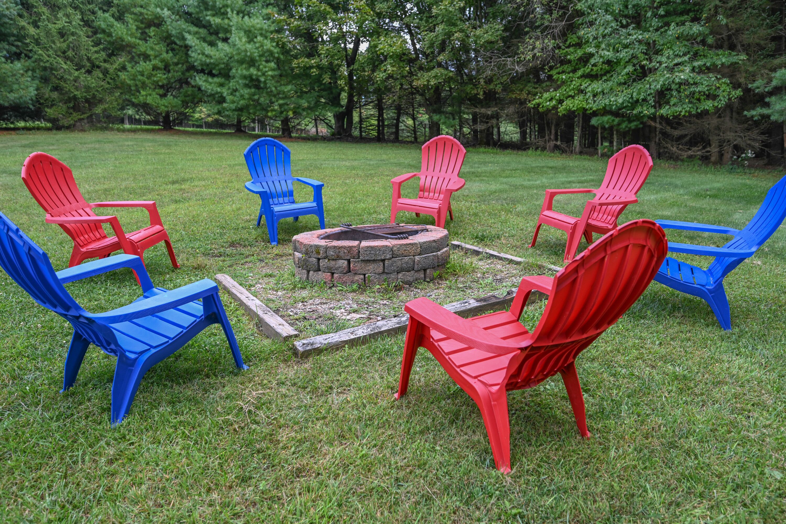 Een cirkel van rode en blauwe Adirondack-stoelen rond een vuurkorf van steen in een gazon.
