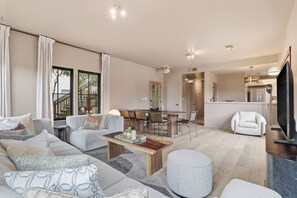 A modern living room with a sofa, coffee table, and ottoman, a dining area with chairs, a kitchen with a TV, and a staircase leading to an upper level.
