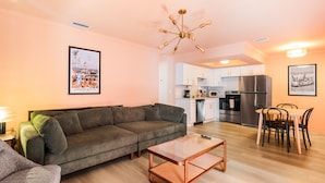A living room with a sofa, a coffee table, a dining table with chairs, a kitchen area with cabinets and appliances, and framed pictures on the walls.