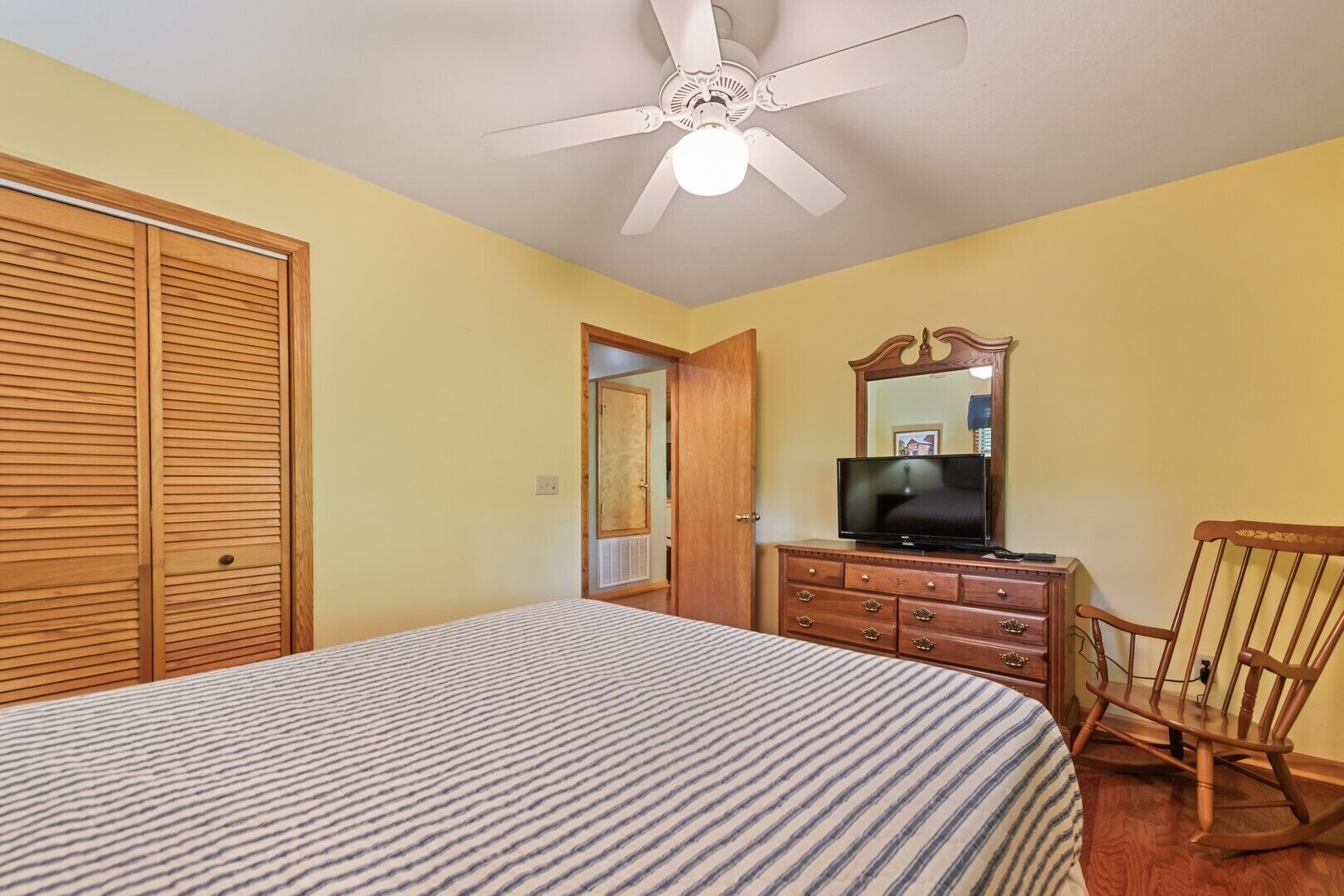 Une chambre à coucher comprenant un lit, une armoire en bois, une commode, un téléviseur et un ventilateur de plafond.