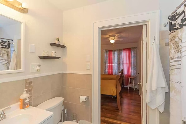A bathroom with a toilet, sink, and shelves. There is a shower curtain with a pattern and a towel hanging on the door. - Yellow Rose House (Amarillo)