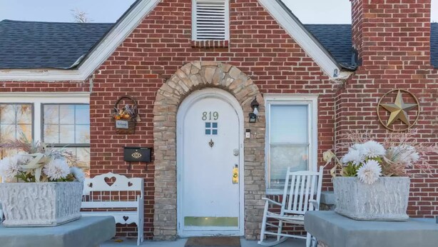 Une maison en briques avec une porte blanche portant le numéro « 819 », un banc blanc et deux jardinières fleuries.