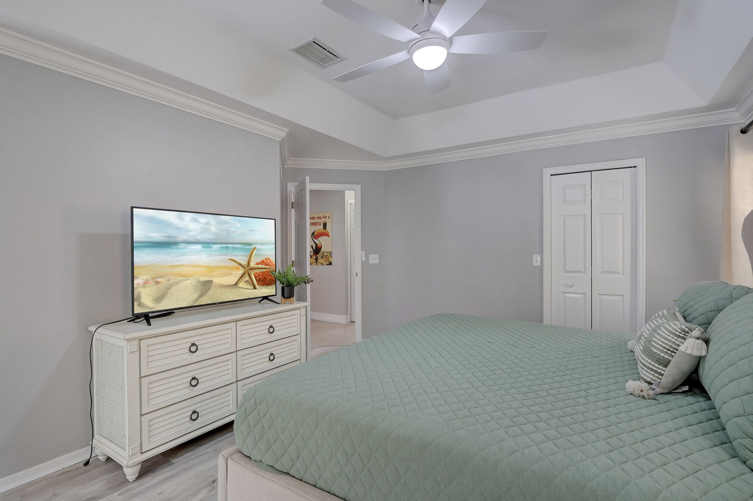 Un dormitorio con cama, televisor sobre una cómoda, ventilador de techo y una puerta.