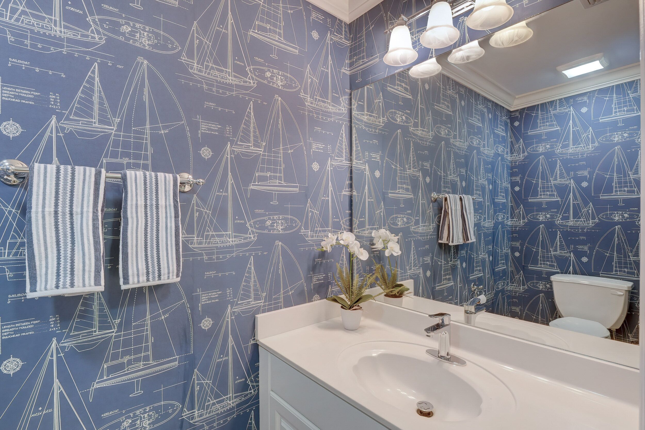 Une salle de bain avec un papier peint sur le thème de la marine, un lavabo blanc et un miroir.