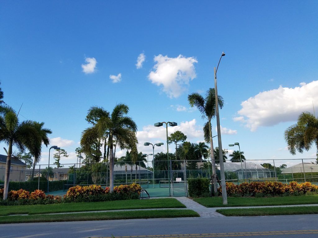 Área externa bem cuidada, com quadra de tênis, palmeiras e um céu azul e límpido.