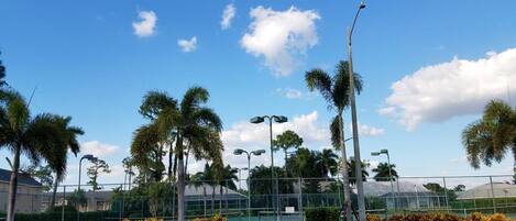 Área externa bem cuidada, com quadra de tênis, palmeiras e um céu azul e límpido.
