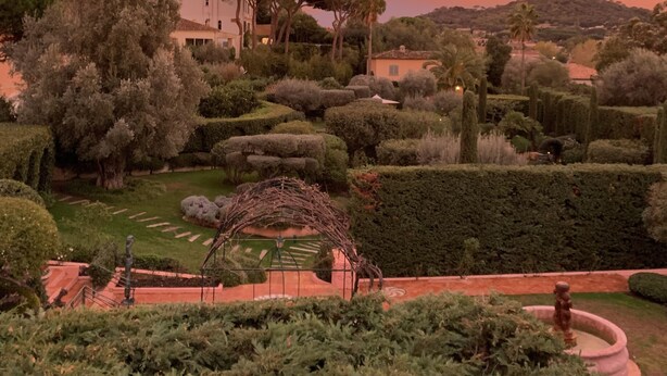 A landscaped garden with neatly trimmed hedges, a fountain, and a building with a red roof.