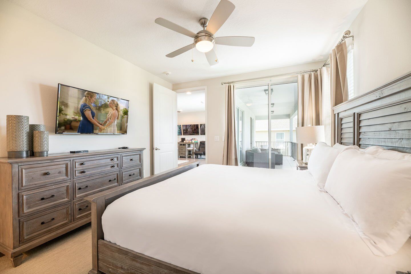 A spacious bedroom with a large bed, a ceiling fan, a TV mounted on the wall, and a view of the balcony through the glass door.
