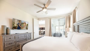 A spacious bedroom with a large bed, a ceiling fan, a TV mounted on the wall, and a view of the balcony through the glass door.