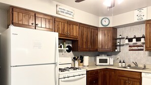 Cucina con mobili in legno, frigorifero bianco, piano cottura, forno a microonde e una sveglia appesa al muro.
