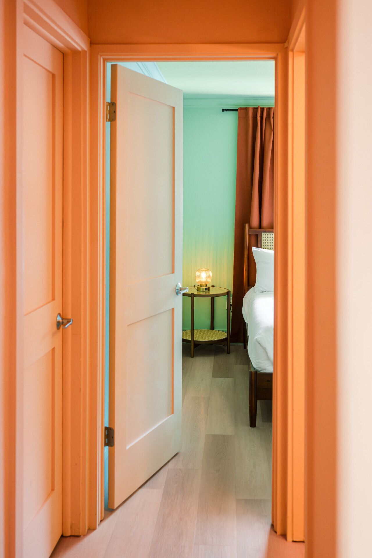 A hotel room with a bed, a bedside table with a lamp, and a view of the hallway.