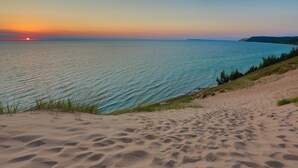 Una playa con arena y agua, un atardecer al fondo y una zona boscosa a la derecha.