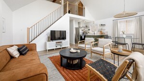 Amplia sala de estar con techo alto, una escalera que conduce a un nivel superior, zona de comedor con muebles modernos y una cocina al fondo.