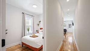 A modern bedroom with a wooden floor, a bed with a white comforter, a bedside table with a lamp, and a window with curtains.