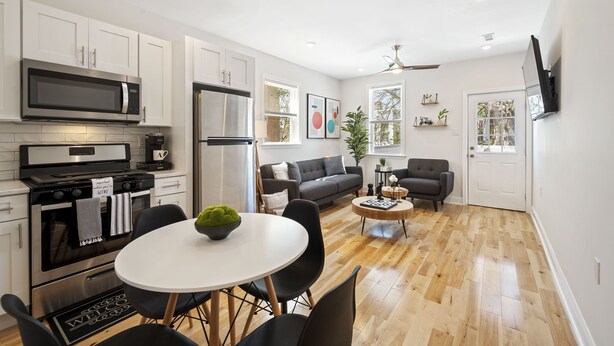 A modern kitchen with white cabinets, a microwave, and a refrigerator. There is a dining area with a round table and chairs, and a living area with a sofa and a coffee table.