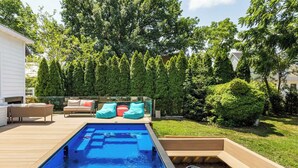 Una piscina rectangular con un borde de madera, rodeada por un deck de madera y una zona verde.