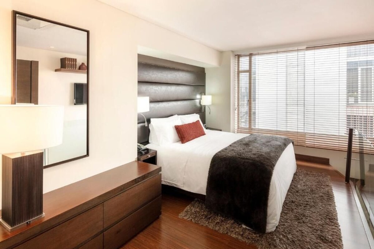 A modern hotel room with a large bed, a nightstand, a dresser, a mirror, and a window with blinds.