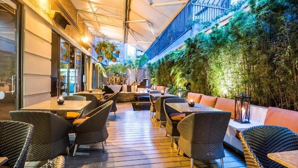 An outdoor cafe with wicker furniture and a wooden deck, surrounded by greenery and a glass wall. - Hotel Cabrera Imperial By Key 33 (Bogotá)