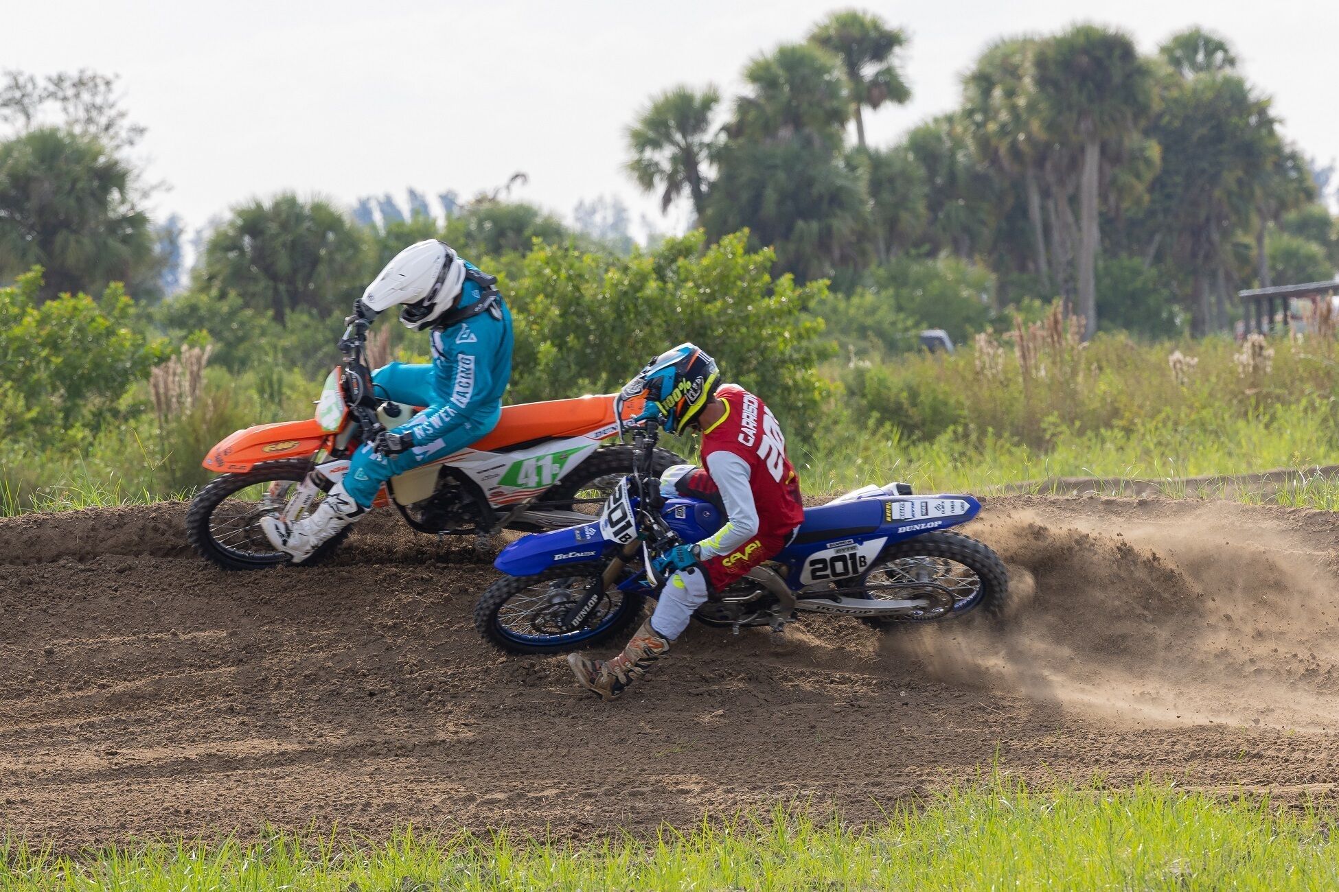 Two dirt bike riders on a track with trees in the background.