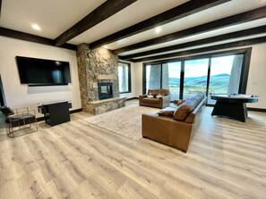 A spacious living room with a stone fireplace, large windows, a flat-screen TV, and a cozy seating area. - Gather on Private Montana Ranch - Sleeps 16 + Incredible Mountain Views, Hot Tub (Belt)