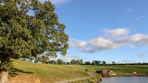 A riverside path with a wooden bridge, a grassy hill, and a small cabin by the water. - Gander Pod (Worcester)