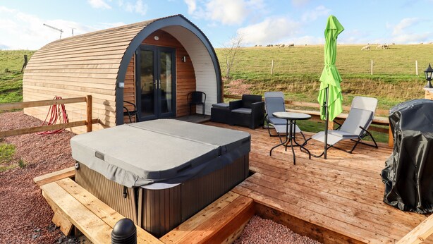 A cozy outdoor hot tub area with a wooden deck, a seating area with chairs and a table, and a small wooden structure with a curved roof. - Gander Pod (Worcester)