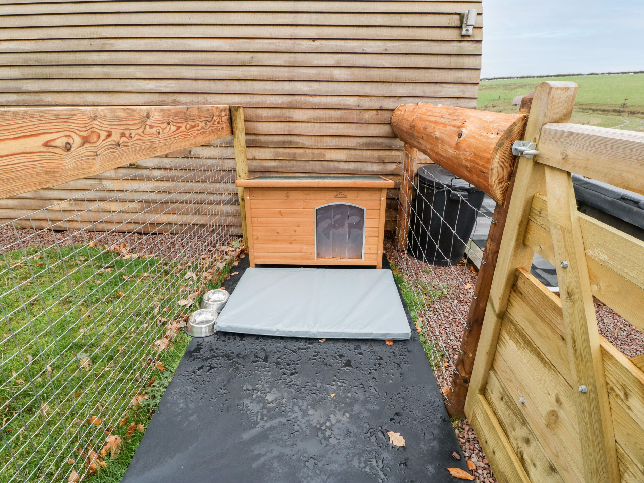 A wooden animal shelter with a fenced entrance, a concrete ramp, and a metal feeding station.