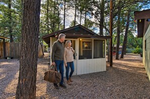 A couple standing in front of a small wooden cabin with a sloped roof, surrounded by trees. - Cabin 2 — Queen Cabin with Sofa Bed and Screened Porch (Lakeside)